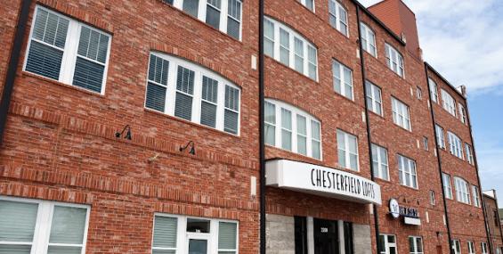 side-angle view of Chesterfield Lofts showcasing its curved brick facade and multiple floors of large, arched windows. The main entrance features a prominent "Chesterfield Lofts" sign above double glass doors. The building's ground-level commercial spaces offer a mix of storefront opportunities in a high-visibility area.