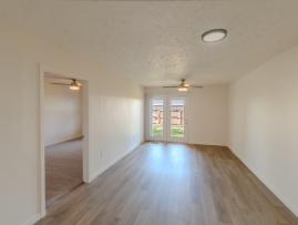 Spacious living room with wood-style flooring, neutral walls, and modern light fixtures. French doors open to a fenced outdoor area, and an adjoining doorway leads into the bedroom.