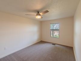 Bright bedroom with neutral walls, new carpet flooring, and a modern ceiling fan with light. A large window provides natural sunlight and overlooks the landscaped outdoor area.