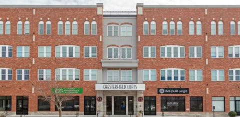 A wide shot of Chesterfield Lofts, highlighting its symmetrical brick architecture with large windows and decorative detailing. The ground floor features multiple businesses, including a yoga studio and wellness center, with additional commercial spaces available for lease. The entrance is centrally located, flanked by retail spaces designed for high foot traffic.
