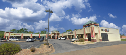 A slightly angled shot of Crossroads Shopping Center, showing various retail stores, including a restaurant with a visible "Hinode" sign. The architectural design features beige and red tones, decorative towers with green rooftops, and a parking lot with multiple vehicles. The sky is partly cloudy with blue patches visible.
